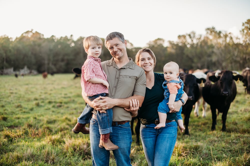 A family photo of two adults and two small children, smiling, with cows and trees in the background. Photo taken at Three Oak Farm. A family photo of two adults and two small children, smiling, with cows and trees in the background. Photo taken at Three Oak Farm.